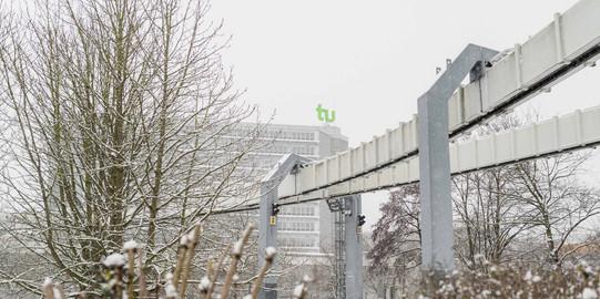 H-Bahn und Mathetower im Winter Kahle Büsche im Vordergrund, im Hintergrund H-Bahn-Strecke und das Mathematikgebäude im Schnee.
