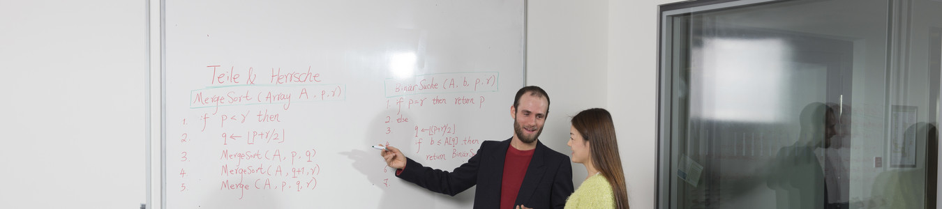 Zwei Personen vor einer Tafel. Eine männliche Person zeigt auf die Tafel und erklärt einer anderen Person etwas.